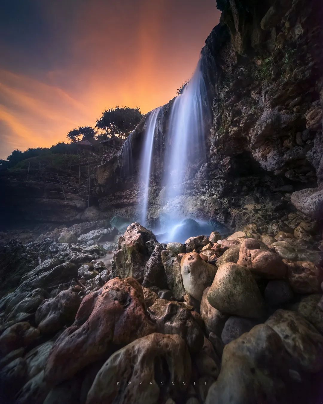 A waterfall at Jogan Beach flowing over rocks with a colorful sunset sky in the background.