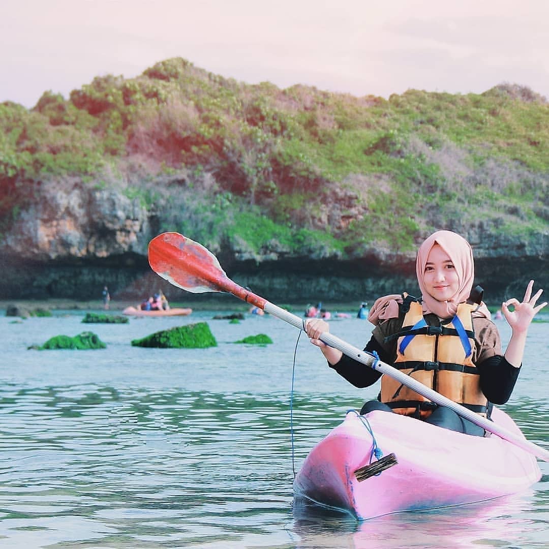 A young woman wearing a hijab and life jacket kayaking on Sadranan Beach, showing an OK gesture.