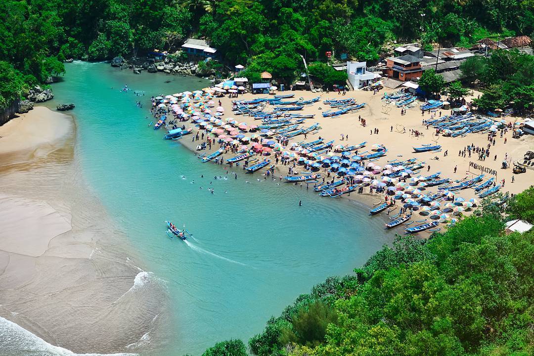 A busy Baron Beach with colorful umbrellas, fishing boats, and visitors swimming in the turquoise water.