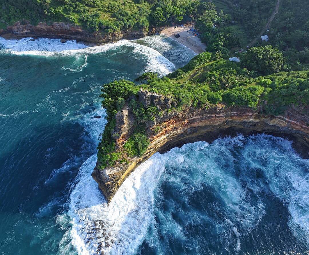 Aerial view of Ngetun Beach with waves crashing against tall cliffs and a sandy shore surrounded by lush greenery.