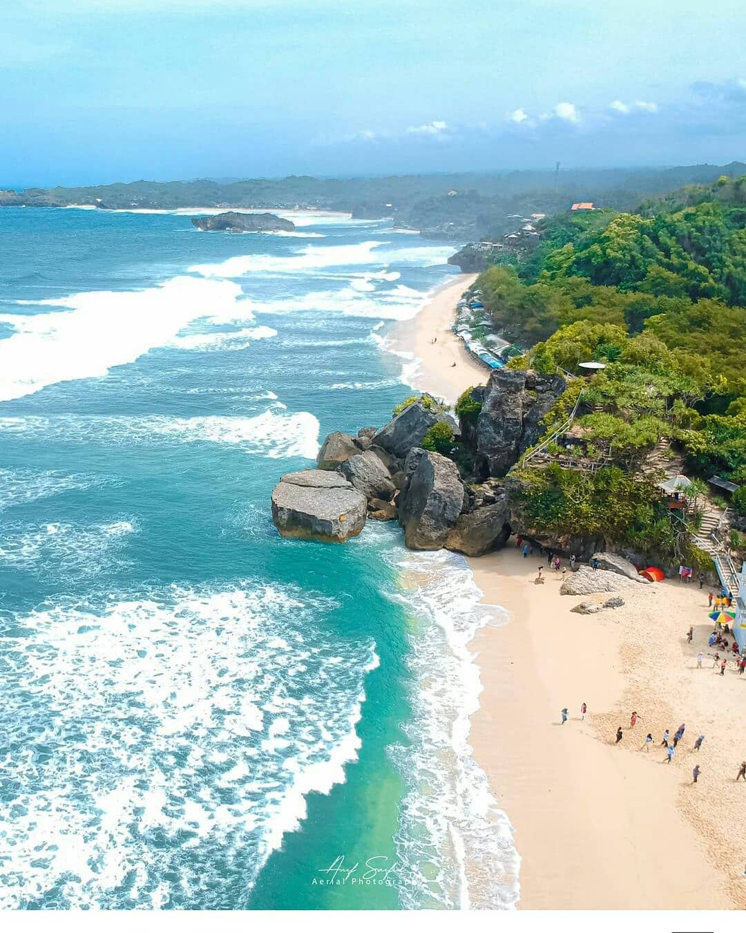 Aerial view of Indrayanti Beach with waves crashing against rocks and lush green hills in the background.