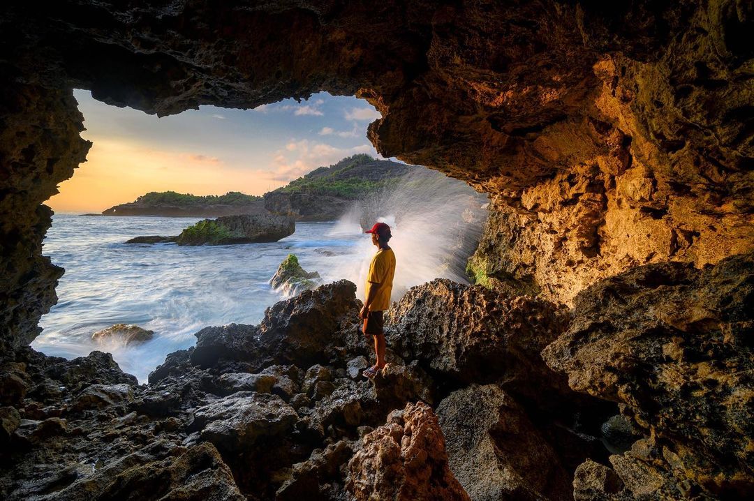 A man standing in a rocky cave, looking out at the ocean waves at Jogan Beach during sunset.