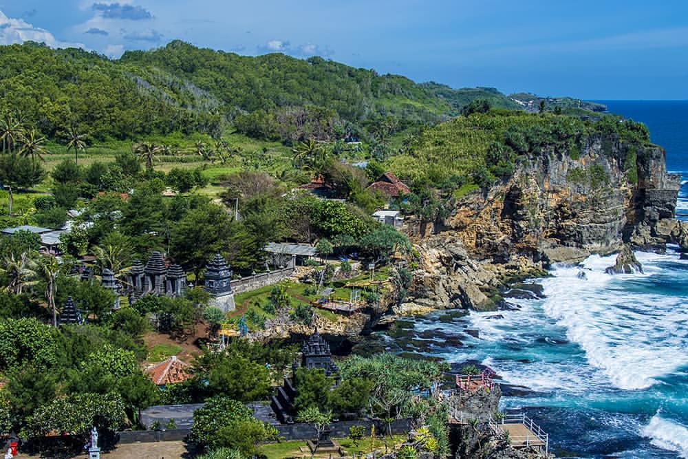 A scenic view of Ngobaran Beach with a Hindu temple and lush greenery on a cliffside.