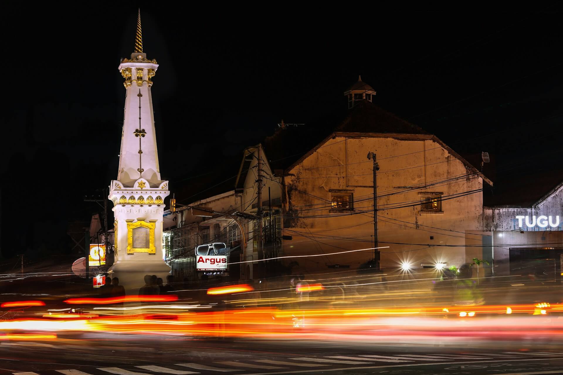 The Tugu Yogyakarta monument is brightly illuminated at night, with light trails from passing vehicles in the foreground.