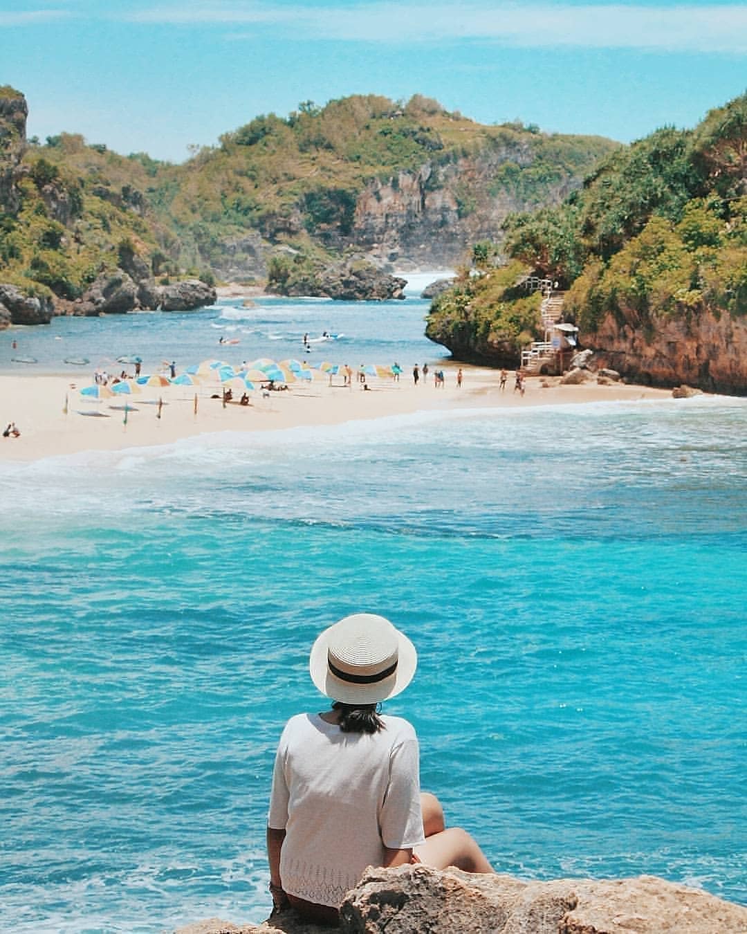 A woman in a white hat sitting on a rock overlooking a turquoise beach with colorful umbrellas and people.