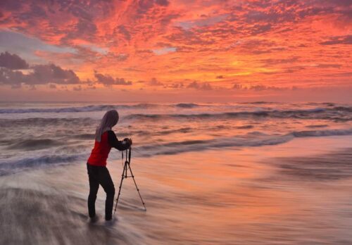 A person standing in the water with a tripod, capturing the sunrise at Trisik Beach.