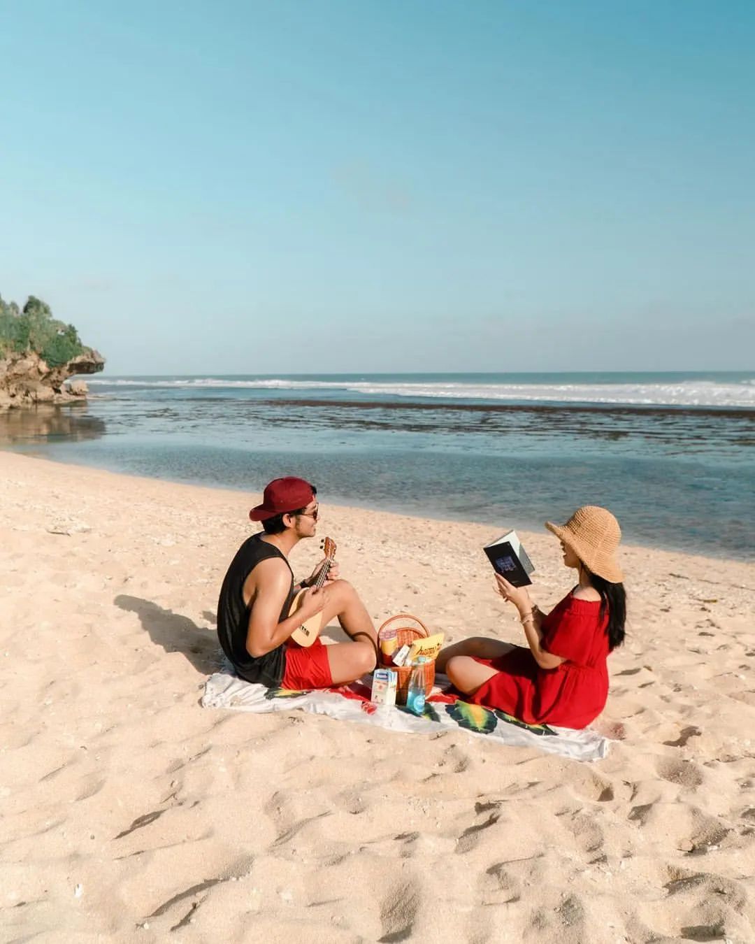 A couple sitting on the sandy beach at Sundak Beach having a picnic, with one playing a ukulele and the other reading a book.