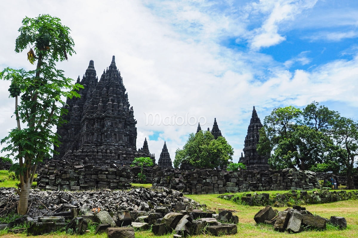 Ruins and trees in front of Prambanan Temple on a partly cloudy day.