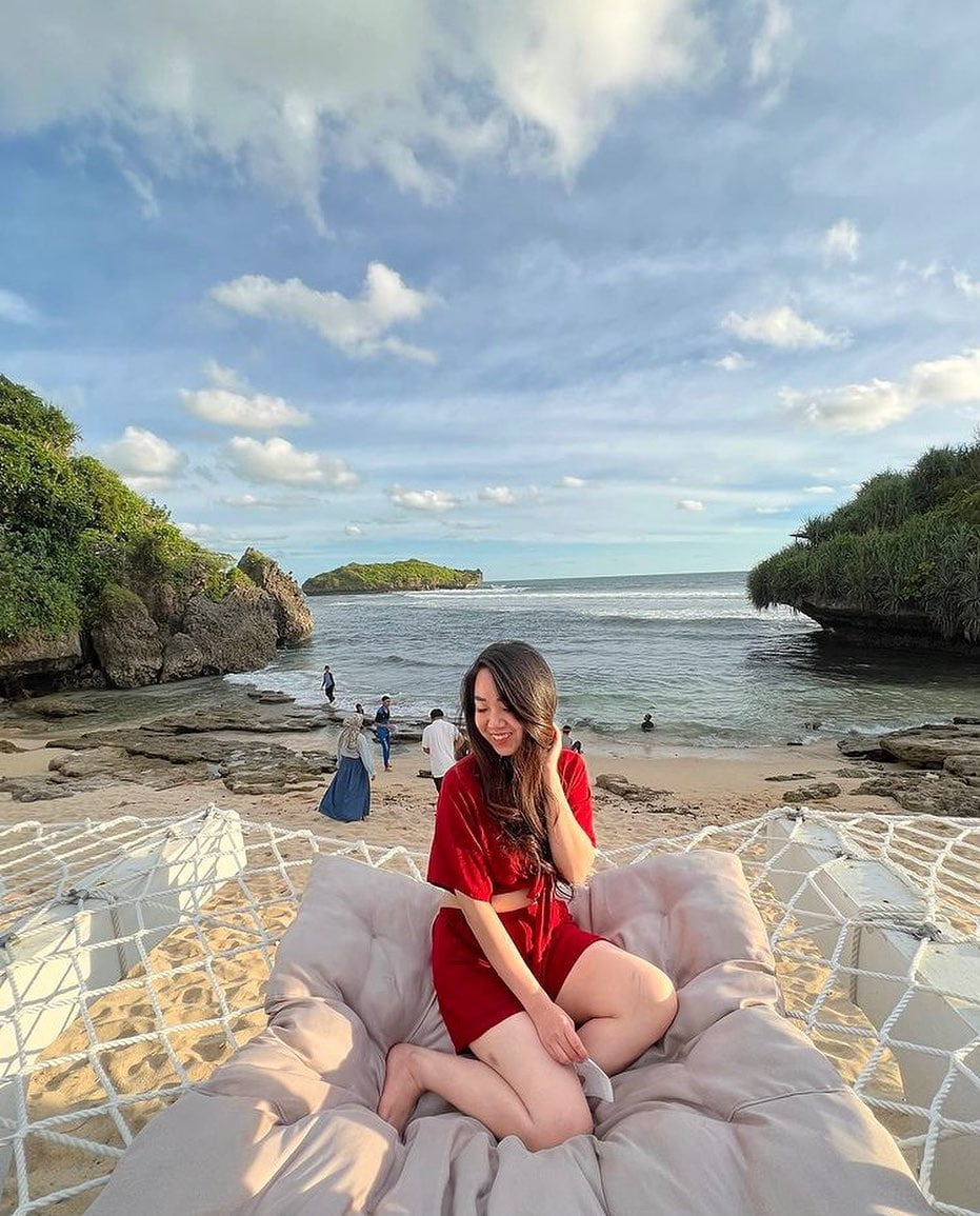 A woman sitting on a hammock bed at Slili Beach with the ocean and cliffs in the background.