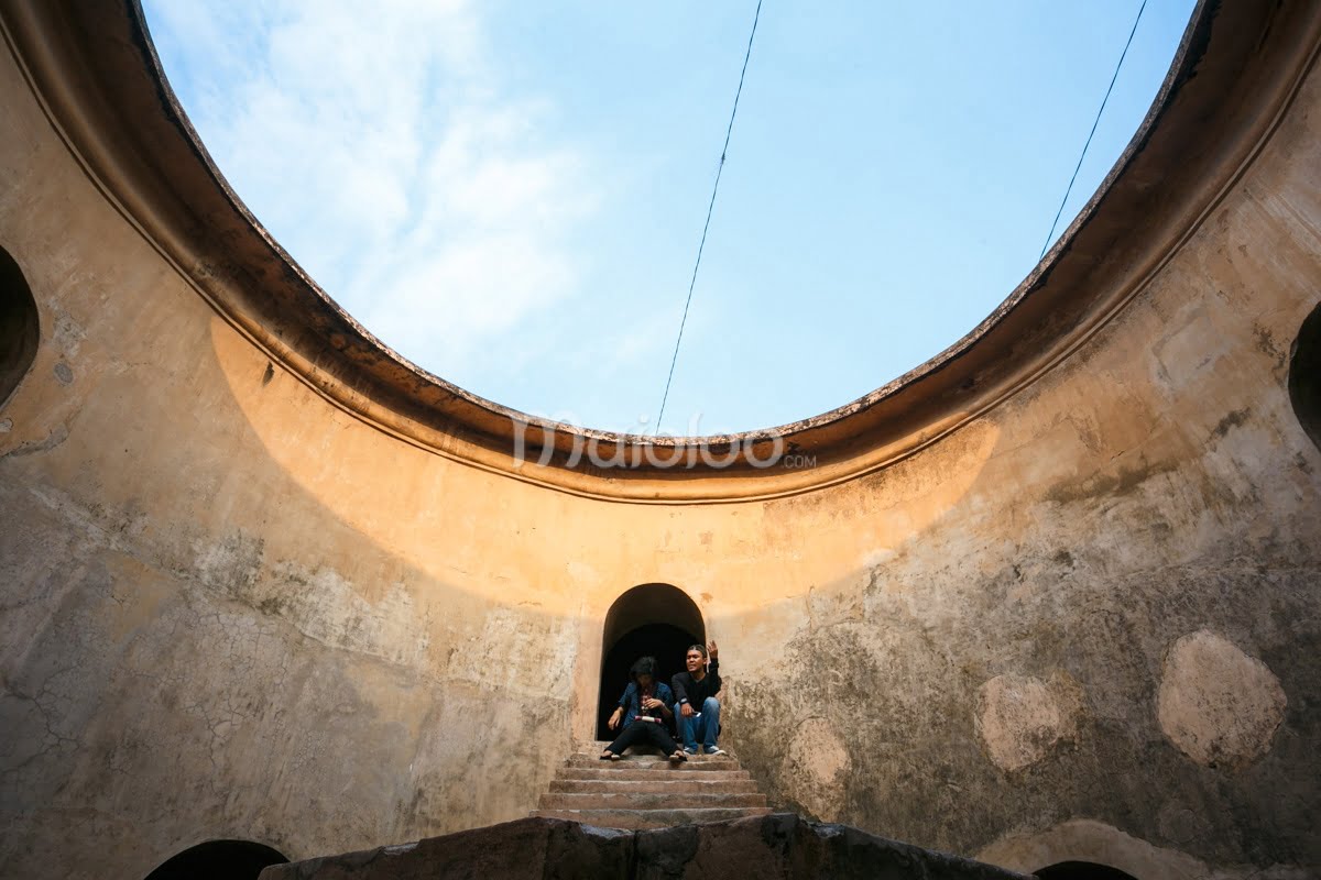 Two visitors sit on steps inside the circular sunlit Sumur Gemuling at Taman Sari, Yogyakarta.