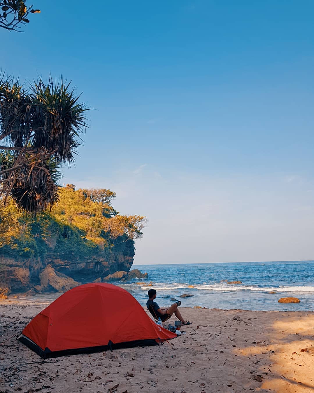 A person sitting on the sandy shore next to a red tent at Ngetun Beach, with cliffs and the sea in the background.