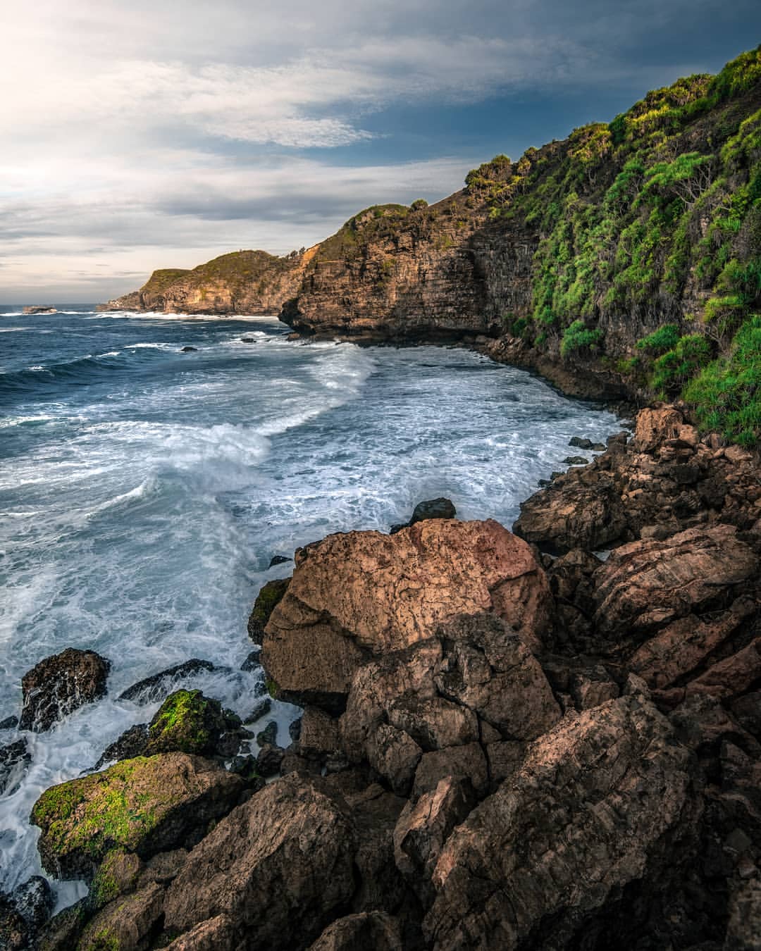 Waves crashing against the rocky cliffs and shore at Ngungap Beach under a partly cloudy sky.