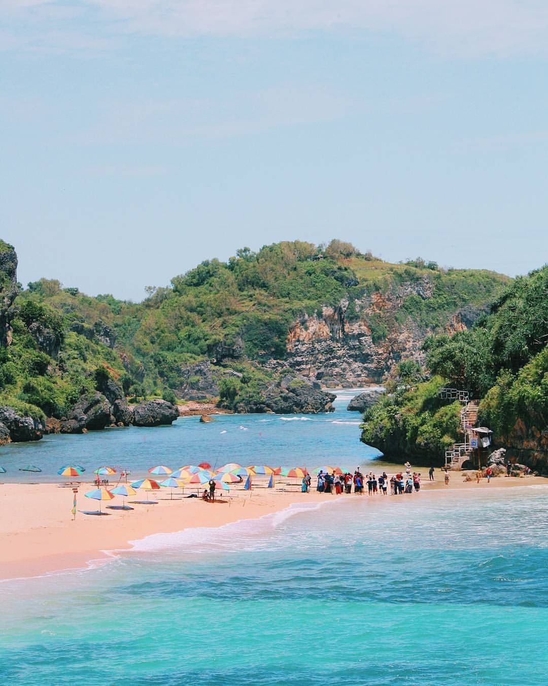 A scenic beach with turquoise water, colorful umbrellas, and green hills in the background.