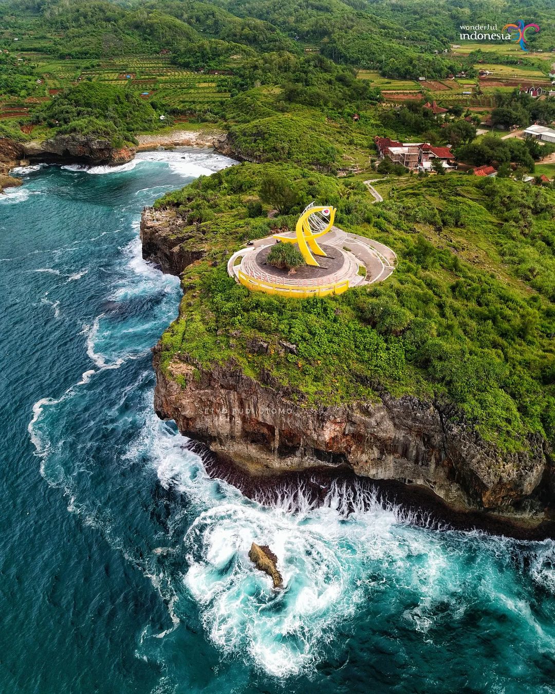 Aerial view of Krakal Beach with its iconic yellow fish statue on a cliffside, surrounded by lush green hills and blue ocean waves.