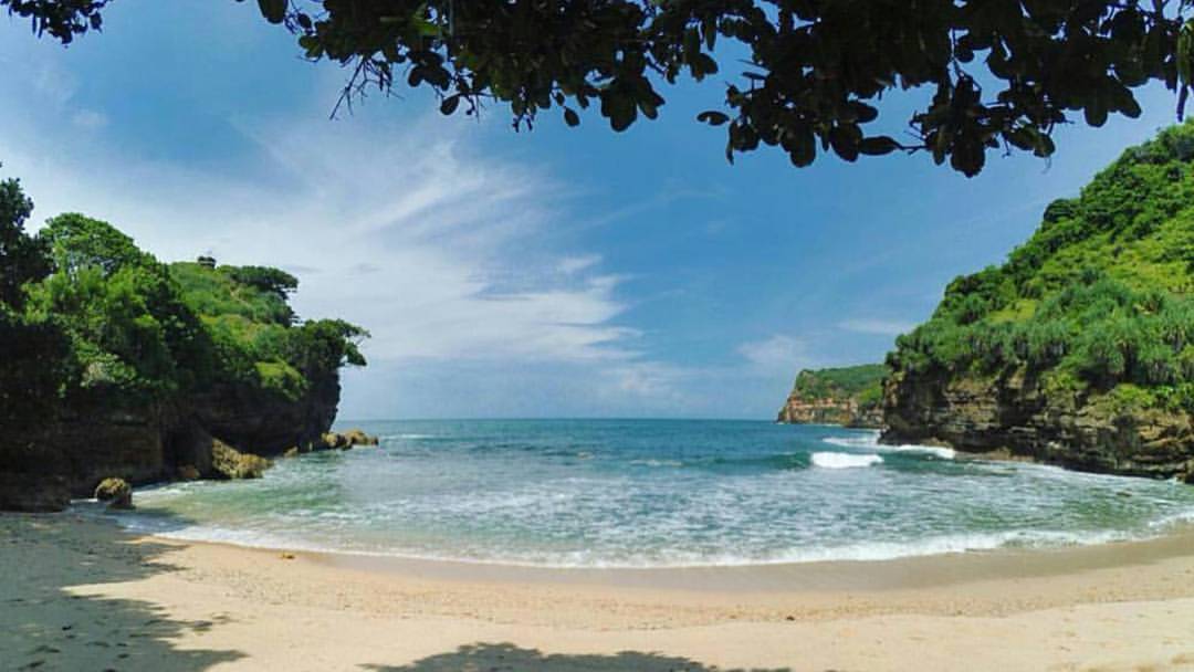 A serene view of the sandy shore at Ngetun Beach with green cliffs on both sides.