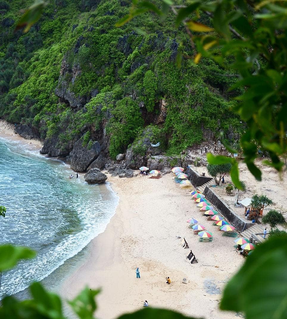 View of Nguyahan Beach with people and colorful umbrellas near green cliffs.