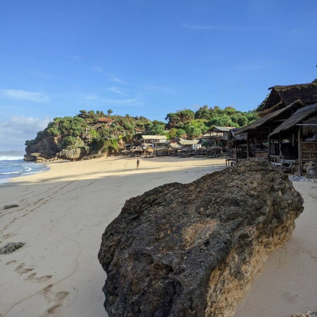 Sandy beach at Watulawang Beach with rocky shore and hillside houses.