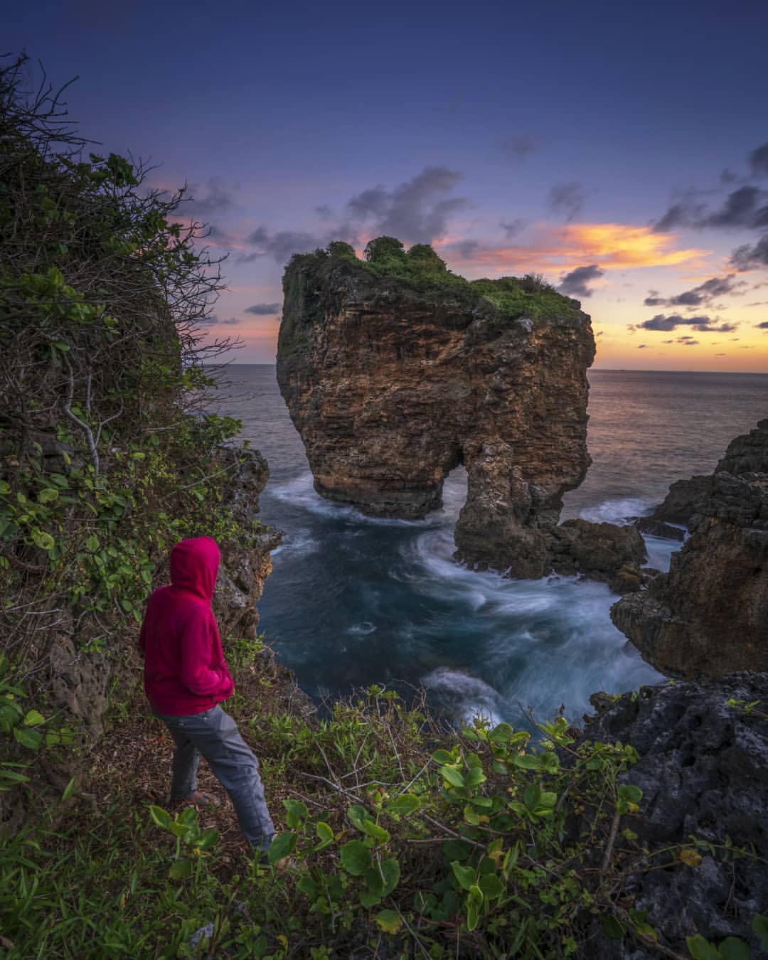 A person in a red hoodie standing on a cliff at Ngungap Beach, overlooking the rock formations and ocean at sunset.