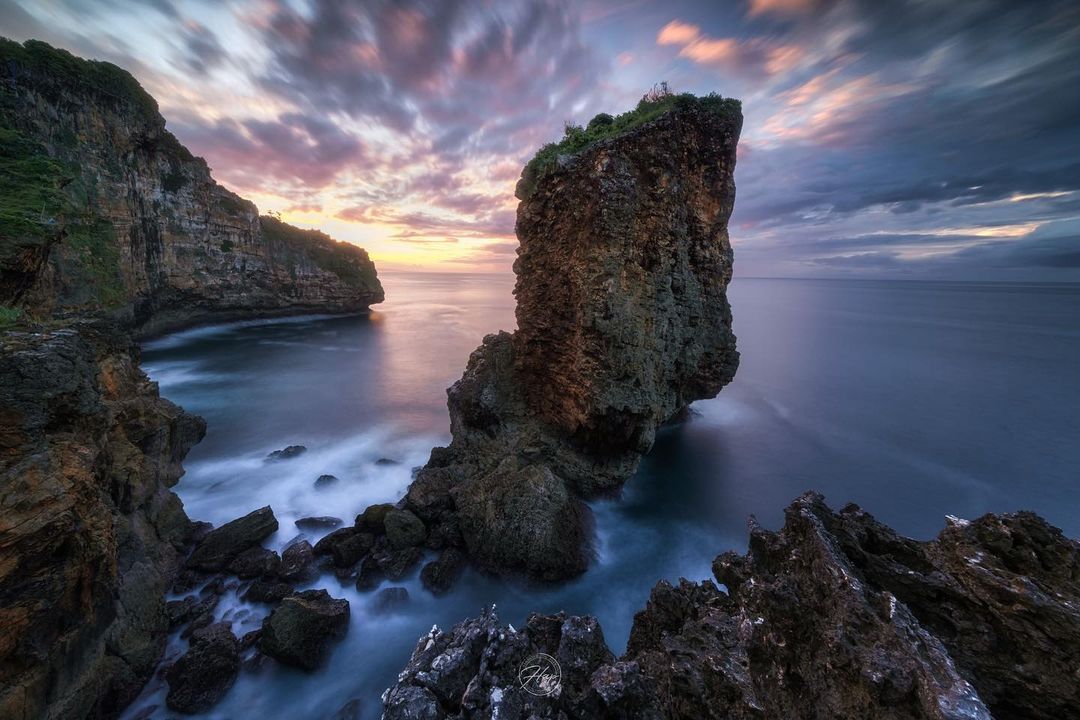 A tall rock formation rising from the sea at sunset at Ngungap Beach with dramatic clouds in the sky.