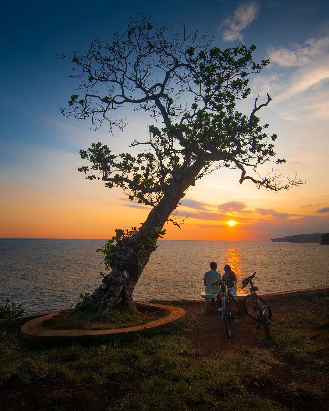 A couple sitting under a tree at Kesirat Beach, watching the sunset with bicycles nearby.