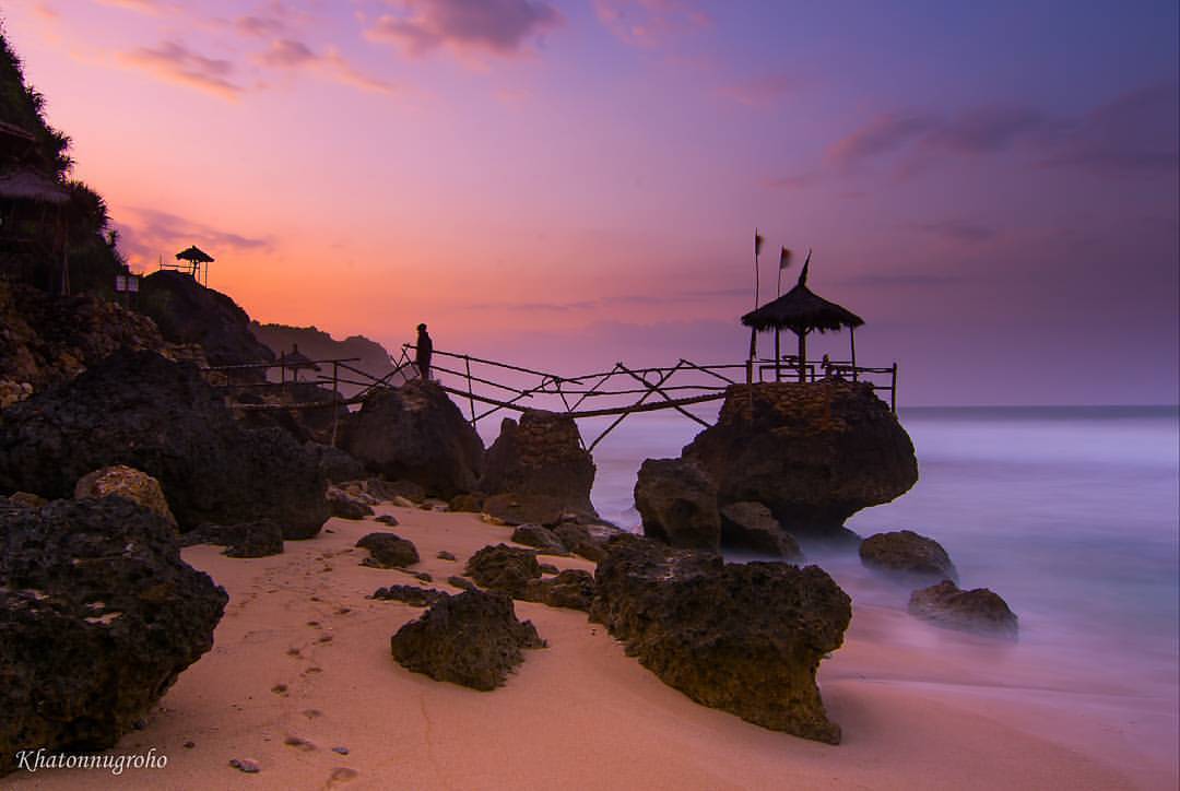 Sunset view at Watulawang Beach with rocky shore and bamboo bridge.