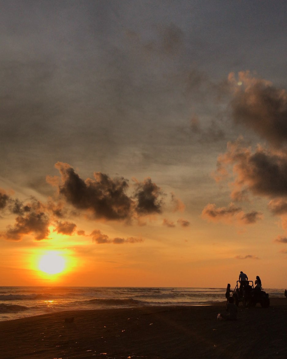 Silhouetted figures and a vehicle on Depok Beach at sunset.