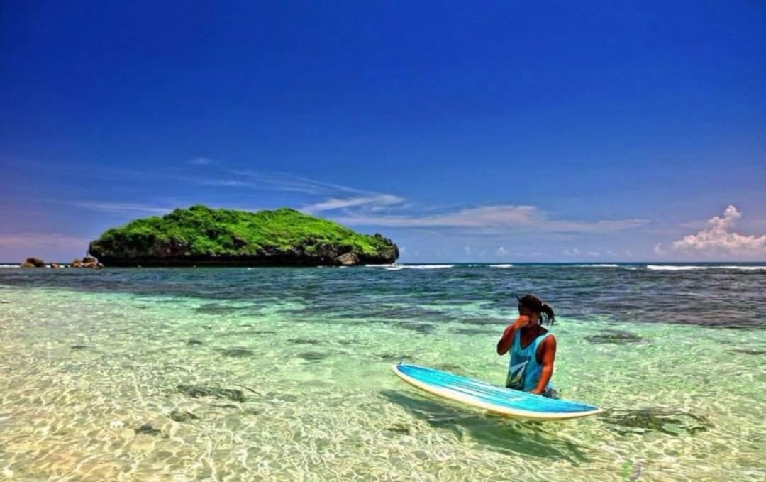 A man stands with a surfboard in the clear shallow waters of Sadranan Beach with a small green island in the background.