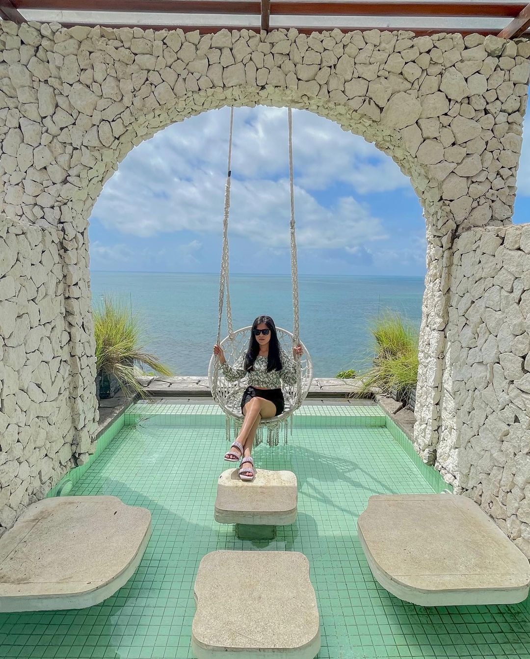 A woman sitting on a swing surrounded by stone arches with an ocean view at HeHa Ocean View.