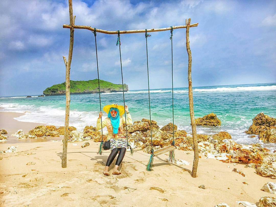 A woman in a blue hijab and yellow hat sits on a swing by the beach at Sadranan Beach with a green island in the background.