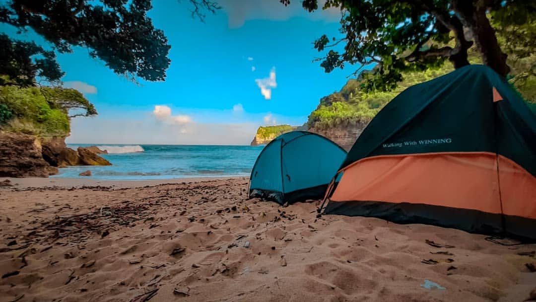 Two tents set up on the sandy shore of Ngetun Beach with the sea and cliffs in the background.