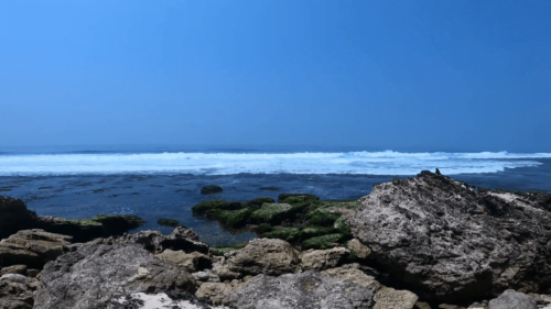 Blue ocean waves crashing against the rocky shore at Kajar Beach under a clear sky.