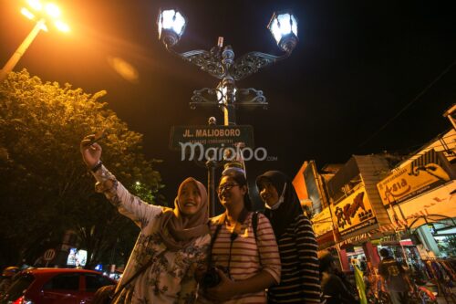 Three tourists taking a selfie under a street sign that reads "Jl. Malioboro" at night.
