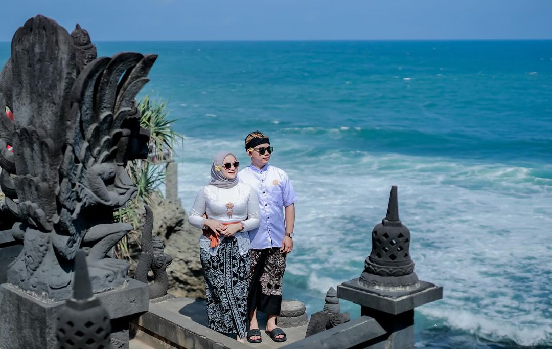 A couple in traditional attire standing near a Hindu statue at Ngobaran Beach with the ocean in the background.