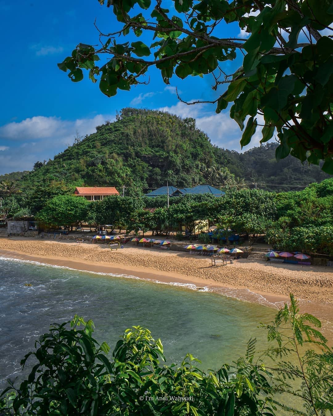 Indrayanti Beach with colorful umbrellas, calm waters, and lush greenery in the background.