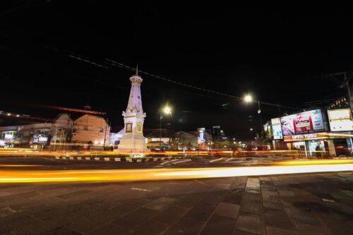 A brightly lit Tugu Yogyakarta stands at a busy intersection at night, with light trails from passing vehicles.