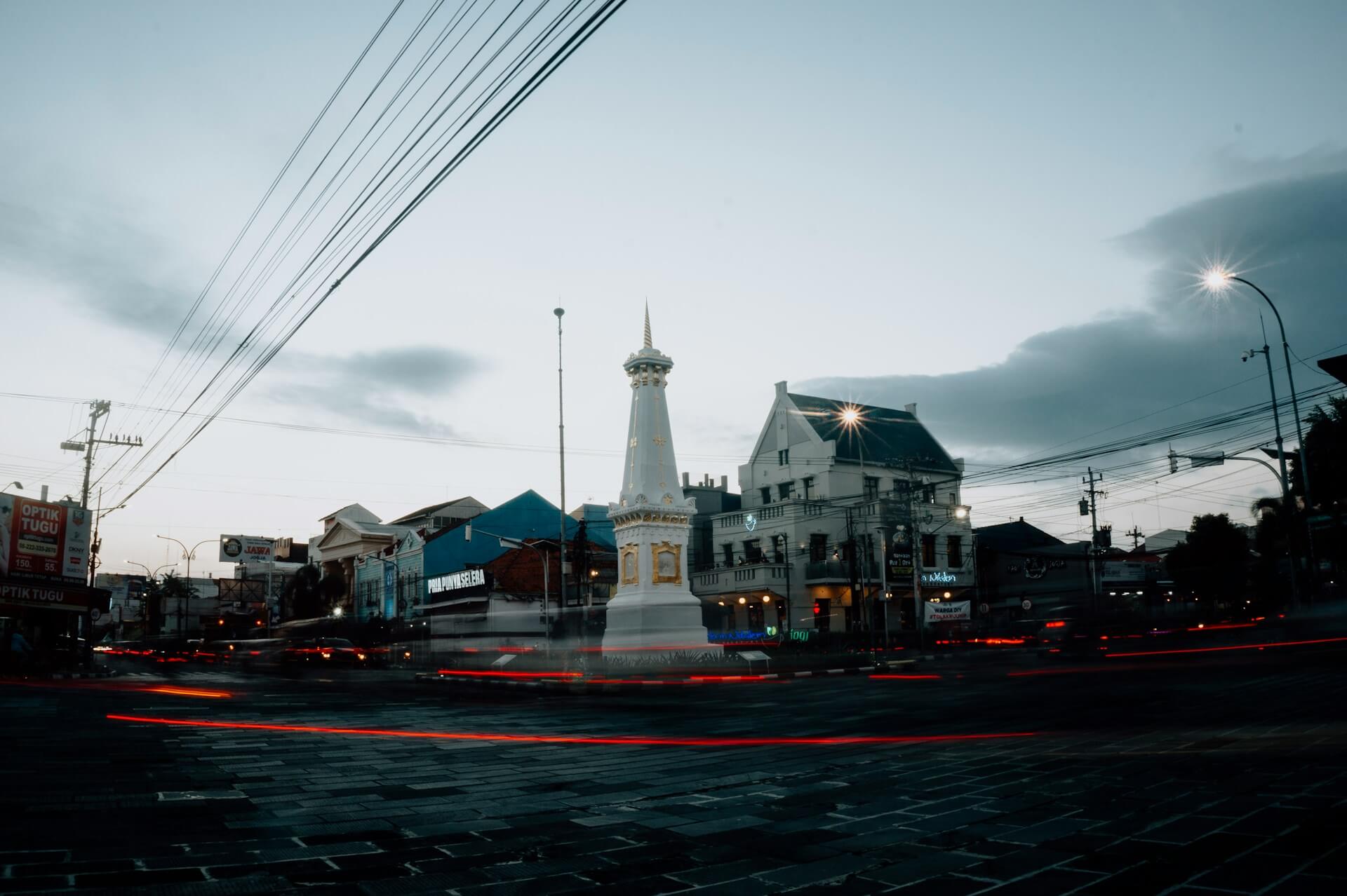 The Tugu Yogyakarta monument stands tall at a busy intersection during dusk, surrounded by buildings and light trails from vehicles.