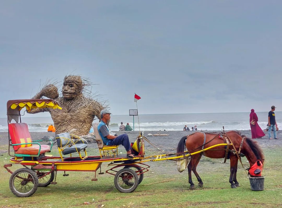 A man sitting in a horse-drawn carriage with a large straw gorilla sculpture in the background at Trisik Beach.