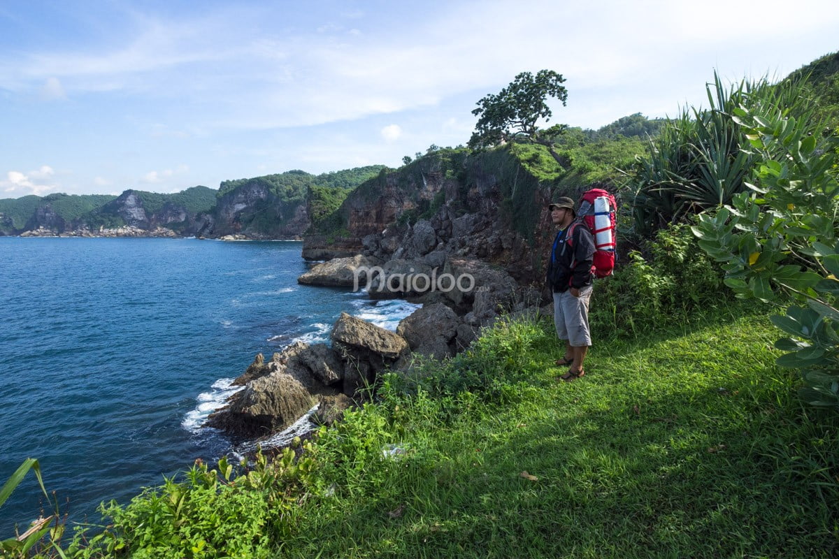 A visitor with a backpack standing on a grassy cliff at Kesirat Beach, looking at the ocean.