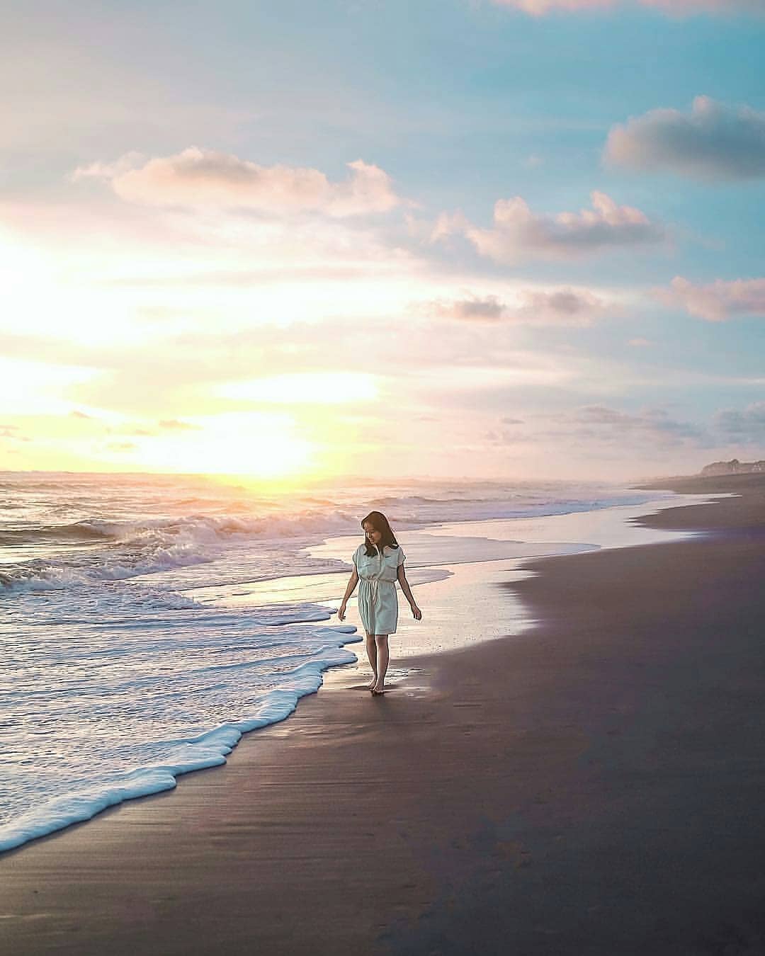 A woman walks along the shore of Depok Beach at sunset.