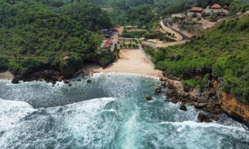 Aerial view of Ngedan Beach in Yogyakarta with clear blue waters, white sandy shores, and surrounding green hills.