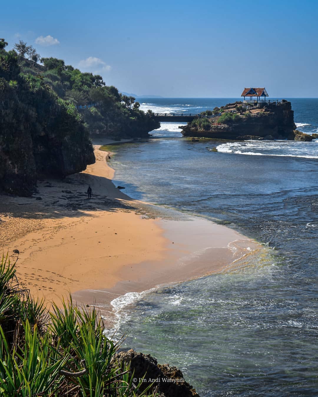 A view of Kukup Beach with its sandy shore, clear waters, and the rocky island pavilion connected by a bridge.