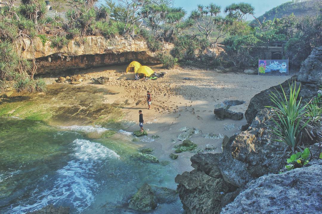 Two people standing near the water on the sandy shore with yellow tents set up at Lolang Beach, surrounded by rocky cliffs and greenery.