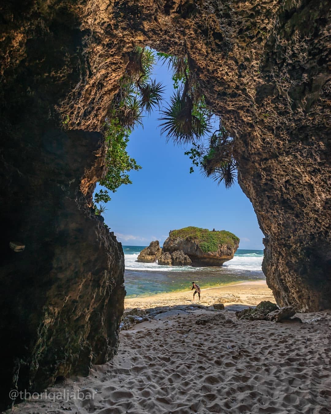 View from inside a cave at Kukup Beach showing a person walking towards the shore and a small rocky island.