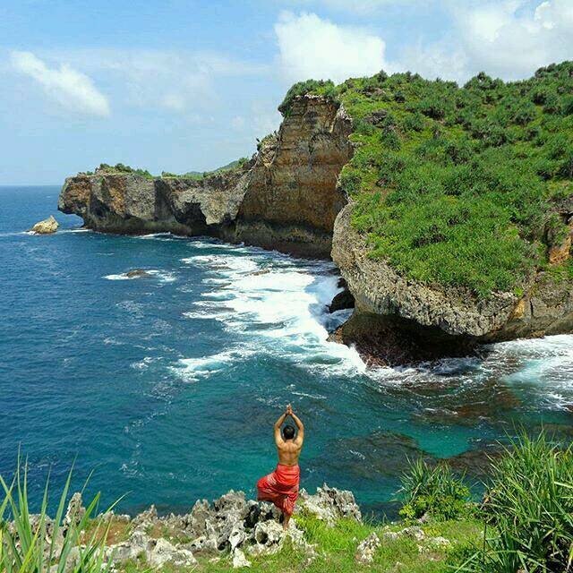 A person standing on a cliff at Ngedan Beach, with arms raised, overlooking the blue ocean and rocky landscape.