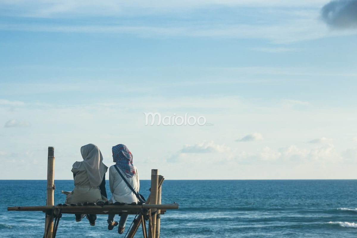 Two visitors sitting on a bamboo platform overlooking the ocean.