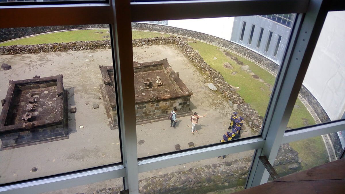 A view through a window of Kimpulan Temple ruins at UII campus in Yogyakarta, featuring stone structures and visitors exploring the site.