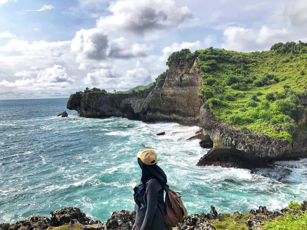 A person wearing a hat and backpack stands on a rocky cliff at Ngedan Beach, overlooking the blue ocean and green hills.