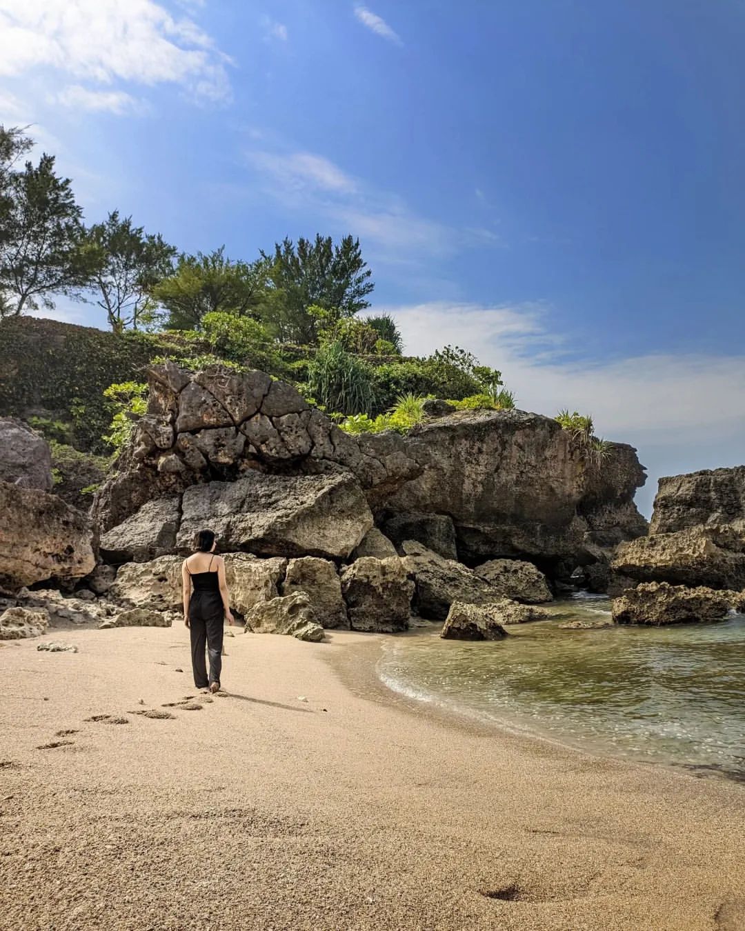 A person walking along the sandy shore with rocky cliffs and greenery in the background at Lolang Beach.