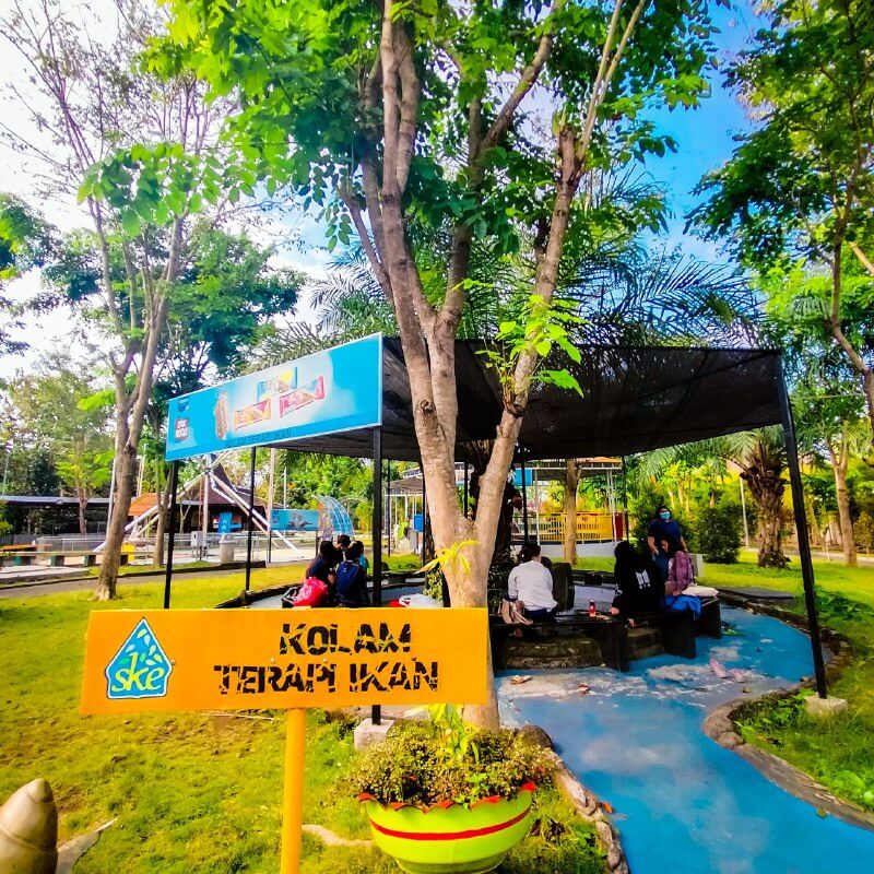 Fish therapy pool area with visitors seated under shaded trees at Sindu Kusuma Edupark.