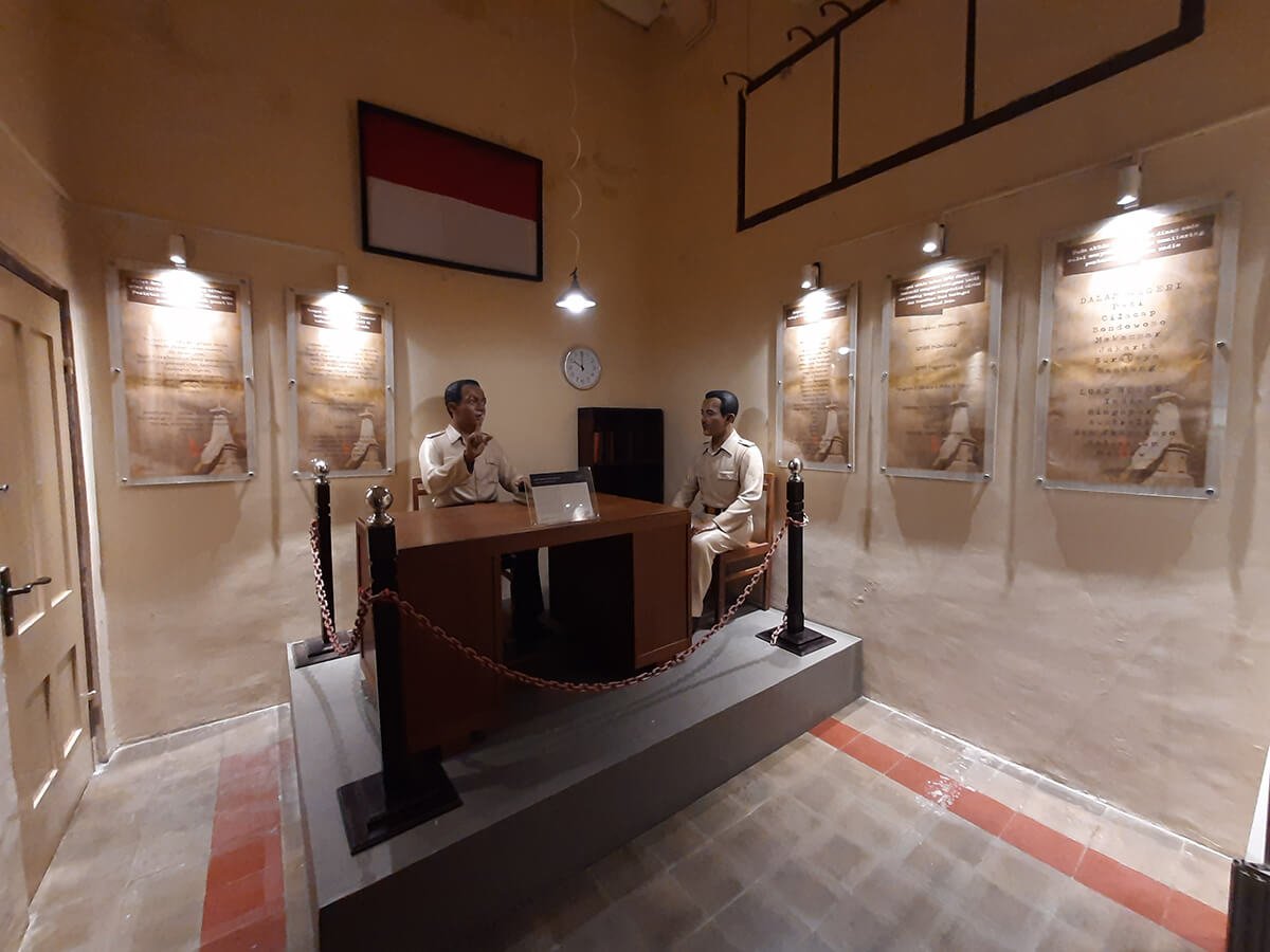 Two mannequins dressed in uniforms sitting at a desk in a museum exhibit featuring historical documents on the walls under display lights.